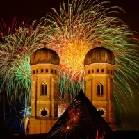 Die Münchner Frauenkirche bei Nacht mit Feuerwerk im Hintergrund.